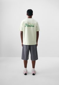 Camiseta de algodón verde claro con "Palmeiras" en letras verdes en la parte trasera, combinada con pantalones cortos de mezclilla grises y zapatillas rosas.
