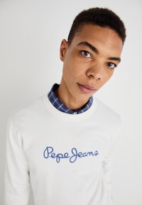 Sudadera blanca con la marca "Pepe Jeans" en azul, superpuesta sobre una camisa de cuadros azul, con cuello redondo y puños acanalados.