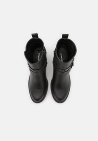 Bottines noires avec une texture en cuir lisse, bout rond, fermeture éclair sur le côté et accents décoratifs en métal. Elles présentent un design à petit talon.
