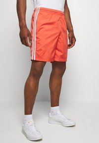 Pantaloni da nuoto arancioni con strisce laterali bianche, dotati di una vita elasticizzata e due tasche laterali. Abbinati a sneaker bianche e calzini.