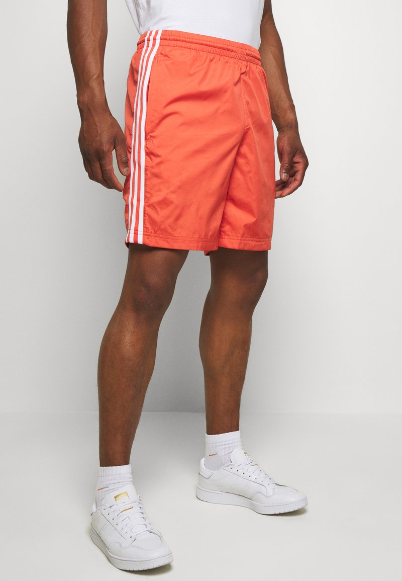 Pantaloni da nuoto arancioni con strisce laterali bianche, dotati di una vita elasticizzata e due tasche laterali. Abbinati a sneaker bianche e calzini.