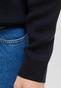 Tuvojies rokas tuvplāns netālu no melna adītu džemperu piedurknes un zilu džinsu aizmugurējās kabatas.