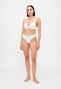 Ensemble de lingerie blanche comprenant un bralette plongeant et un string assorti. Le bralette possède des bretelles fines, et le string a une taille texturée avec le logo "HUGO".