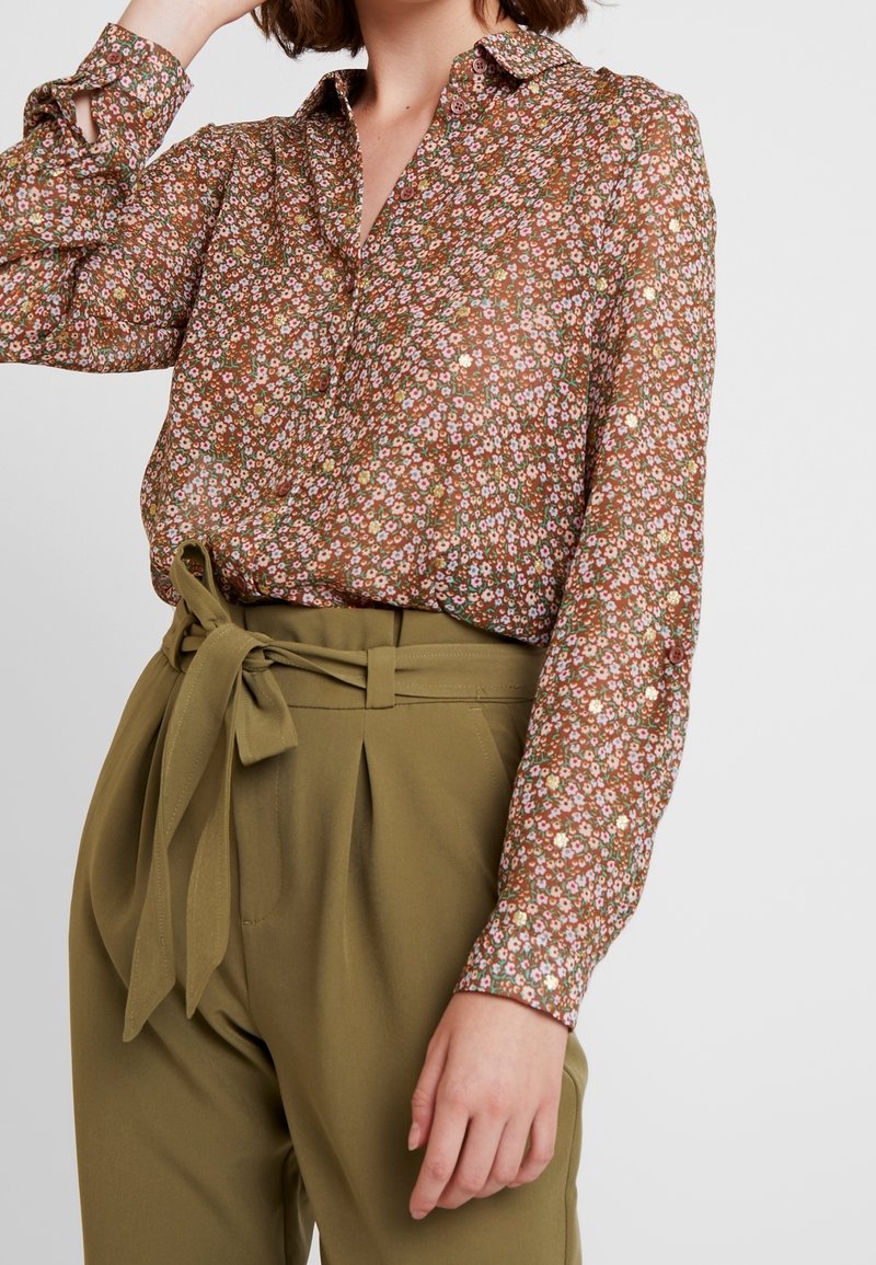 Brun blommig blus med långa ärmar, med en knapp-up krage och en lätt textur, kombinerad med högmidjade olivgröna byxor.