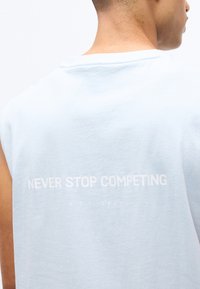 Άνδρας που φορά ένα ανοιχτό μπλε αμάνικο μπλουζάκι με την επιγραφή «NEVER STOP COMPETING EST 2017» στην πλάτη, πάνω σε λευκό φόντο.