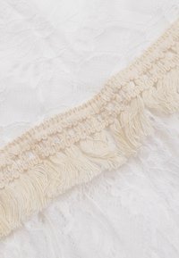 Tessuto di pizzo bianco con motivi floreali, accentuato da un bordo frangiato color crema lungo il margine, che mette in risalto la texture e i dettagli.