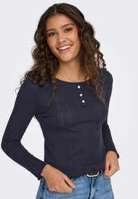 Marineblå langermet topp laget av teksturert stoff, med rund hals, lukkes med tre knapper, og frynsete kant.