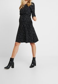 Kobieta w czarnej, długiej sukience ciążowej w białe kropki, z czarnymi botkami do kostek, pozująca na tle jednolitego, białego tła.