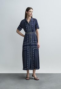 Γυναίκα που στέκεται με το ένα χέρι στη μέση, φορώντας μια μίντι φόρεμα με ναυτικό μπλε σχέδιο και μαύρα σανδάλια με τακούνι, μπροστά σε απλό φόντο.