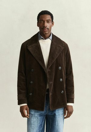 Mann trägt dunkelbraunen Cord-Doppelreihermantel, weißen Strickpullover, blaue Jeans und steht vor einem schlichten beigen Hintergrund.