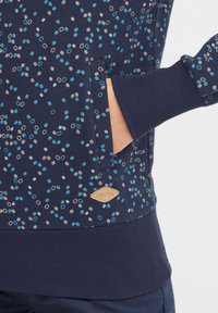 Marinblå sweatshirt med flerfärgad prickmönster, ribbade ärmslut, fickdetalj och en beige etikett med "oxmo" vid nederkanten.