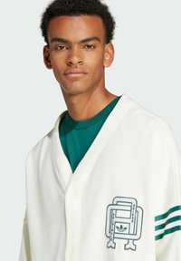 Vit cardigan med v-ringning, med en grön broderad logotyp på bröstet och randiga detaljer på ärmarna; tillverkad av mjukt tyg.