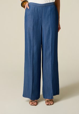 Femme portant un pantalon en jean bleu taille haute à jambes larges et des sandales à bout ouvert avec des brides à motif serpent, debout avec la main dans la poche.