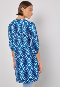 Blaue und weiße gemusterte Tunika mit Dreiviertelärmel, locker sitzend und mit einem längeren Rückensaum, aus leichtem Material gefertigt.