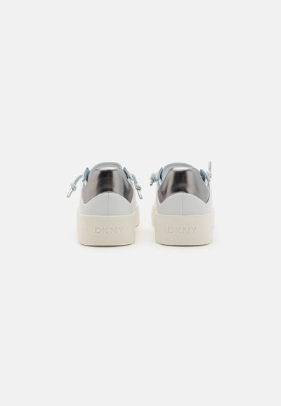 Baskets plates blanches avec un dos argent métallique et des accents bleus clairs, dotées d'une semelle en caoutchouc texturée et du logo DKNY sur le talon.