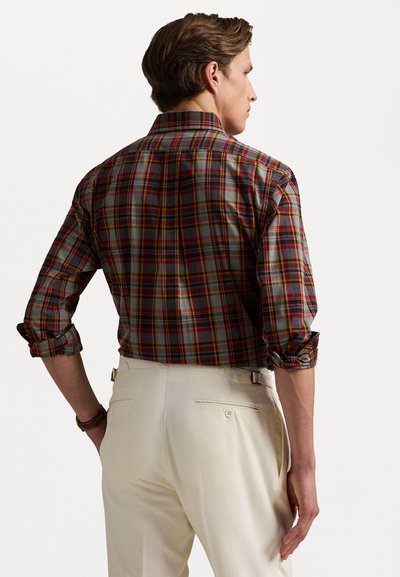 Chemise à carreaux avec des carrés multicolores en rouge, vert et gris. Manches longues retroussées, portée avec un pantalon de couleur crème. Vue arrière affichée.