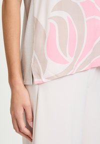 Luźna bluzka z lekkiego materiału, ozdobiona dużym wzorem kwiatowym w odcieniach różu i beżu, z delikatnie teksturowanym wykończeniem i bocznym rozcięciem.