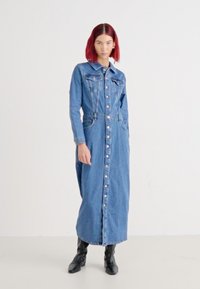 Denim maksi-kjole med knappelukking foran, spiss krage, to brystlommer, lange ermer og en avslappet passform. Brukt med svarte ankelstøvler.