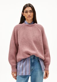 Strickpullover in staubigem Pink mit geripptem Muster und überschnittenen Schultern, kombiniert mit einem hellblauen gestreiften Hemd und Jeans.