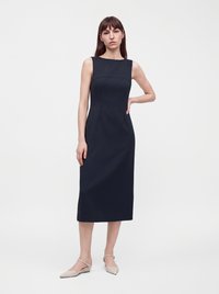 Femme portant une robe midi noire sans manches et des ballerines beiges pointues, debout contre un fond clair uni.