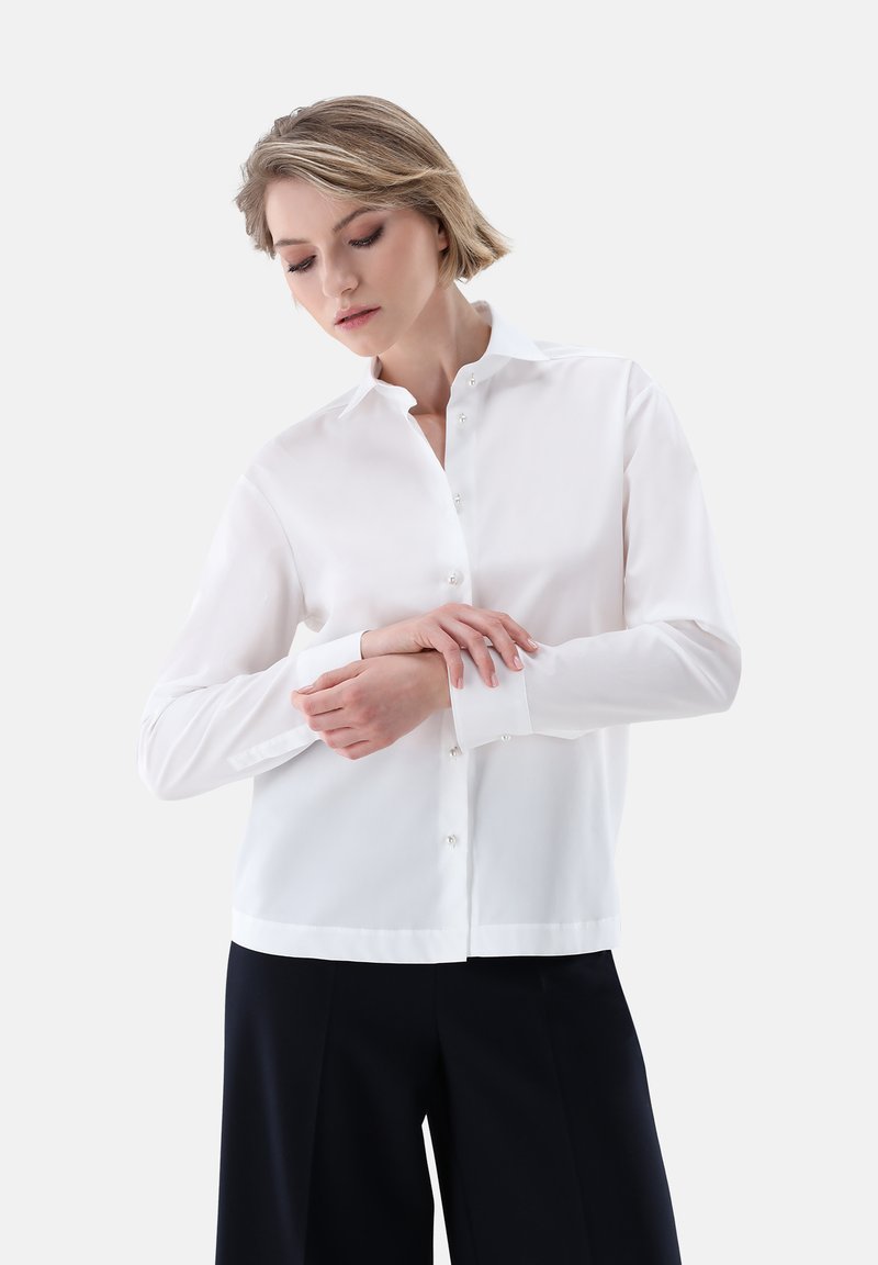 Camicia bianca a maniche lunghe con colletto aderente e taglio dritto. Tessuto morbido, polsini con un bottone e leggera lucentezza.