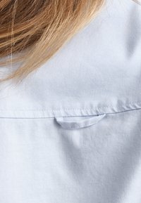 Světle modrá tkanina košile s texturovaným povrchem, s malou smyčkou na zadní straně u límce pro zavěšení nebo úpravu.