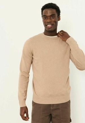 Lächelnder Mann, der den Kragen eines beigen Pullovers richtet, braune Hose trägt und vor einem einfarbigen hellen Hintergrund steht.