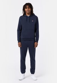 Námořnická modrá mikina s kapucí a odpovídající joggingové kalhoty, obsahující přední kapsu, kapuci se stahovací šňůrkou a elastické manžety. Hladká látka bez viditelných vzorů.