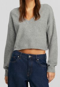 Sueldo gris, ajustado, de punto con escote en V y puños acanalados, combinado con pantalones vaqueros de mezclilla azul oscuro que presentan una cintura ajustada y costuras en contraste.