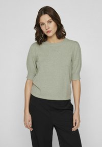 Pull en tricot vert clair avec des manches courtes bouffantes, un col rond et une coupe courte, associé à un pantalon noir sur un fond uni.