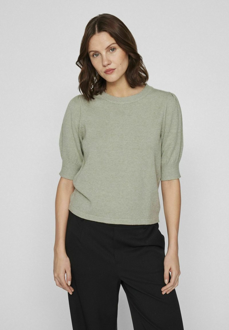 Pull en tricot vert clair avec des manches courtes bouffantes, un col rond et une coupe courte, associé à un pantalon noir sur un fond uni.