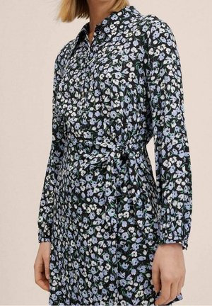 Kobieta ubrana w czarną sukienkę z długim rękawem, z jasnoniebieskim i białym kwiatowym wzorem, z detalem wiązanym w talii.