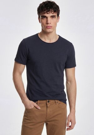 Junger Mann mit kurzen lockigen Haaren, der ein dunkelblaues T-Shirt und braune Hosen trägt, steht mit einer Hand in der Hosentasche vor einem schlichten Hintergrund.