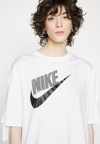 Camiseta blanca de manga corta con un prominente logo negro de Nike, texto a rayas y un diseño de swoosh elegante. Material de algodón, ajuste relajado.