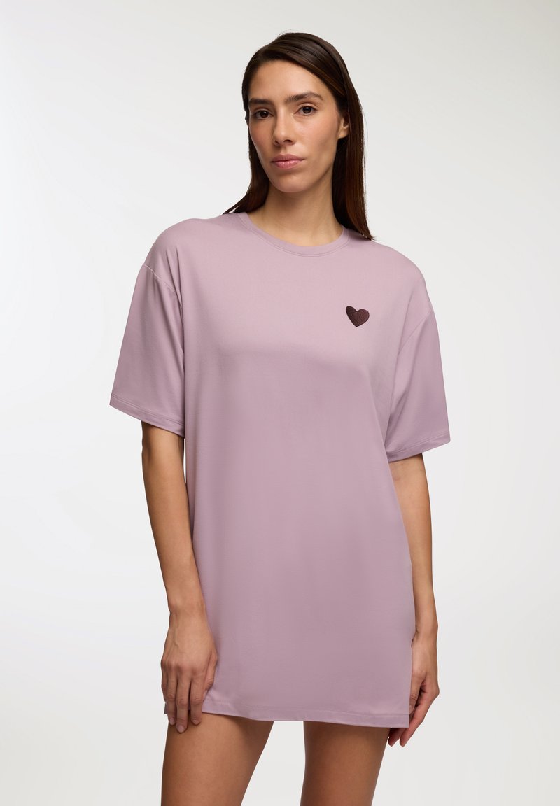 Maglietta oversize rosa con maniche corte e un piccolo design di cuore marrone sul lato sinistro. Tessuto morbido con una superficie liscia.