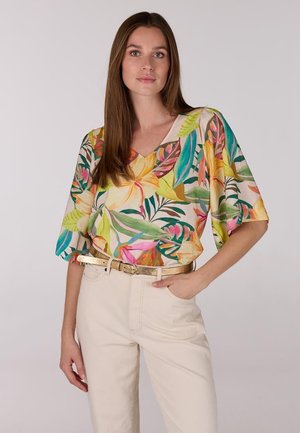 Bloemenprint blouse met wijde mouwen, in levendige kleuren zoals roze, geel en groen op een lichte achtergrond, gedragen met lichtbeige broek en een gouden riem.