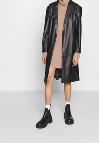 Manteau en faux cuir noir avec un revers structuré, des manches longues et une coupe décontractée, associé à une robe en tricot beige et des bottines noires épaisses.