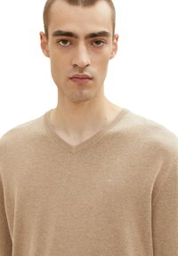 Beige V-Ausschnitt-Pullover aus weichem Strickmaterial, mit dezentem Logo-Detail auf der linken Brust und einem lässigen Schnitt.