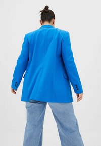 Blazer oversize bleu vif avec épaules structurées, fermeture à un bouton et boutons noirs, associé à un jean large bleu clair.