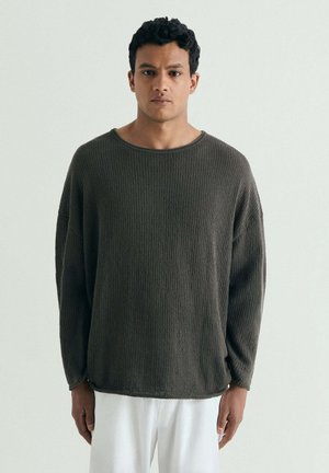 Joven con pelo corto y rizado, que lleva un suéter de punto holgado gris oscuro y pantalones blancos, de pie frente a un fondo liso.