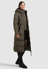 Lang, fluffy olivengrøn parka med hætte, sorte lynlåslommer og en glat tekstur. Sammen med en sort sweater og ankelstøvler.