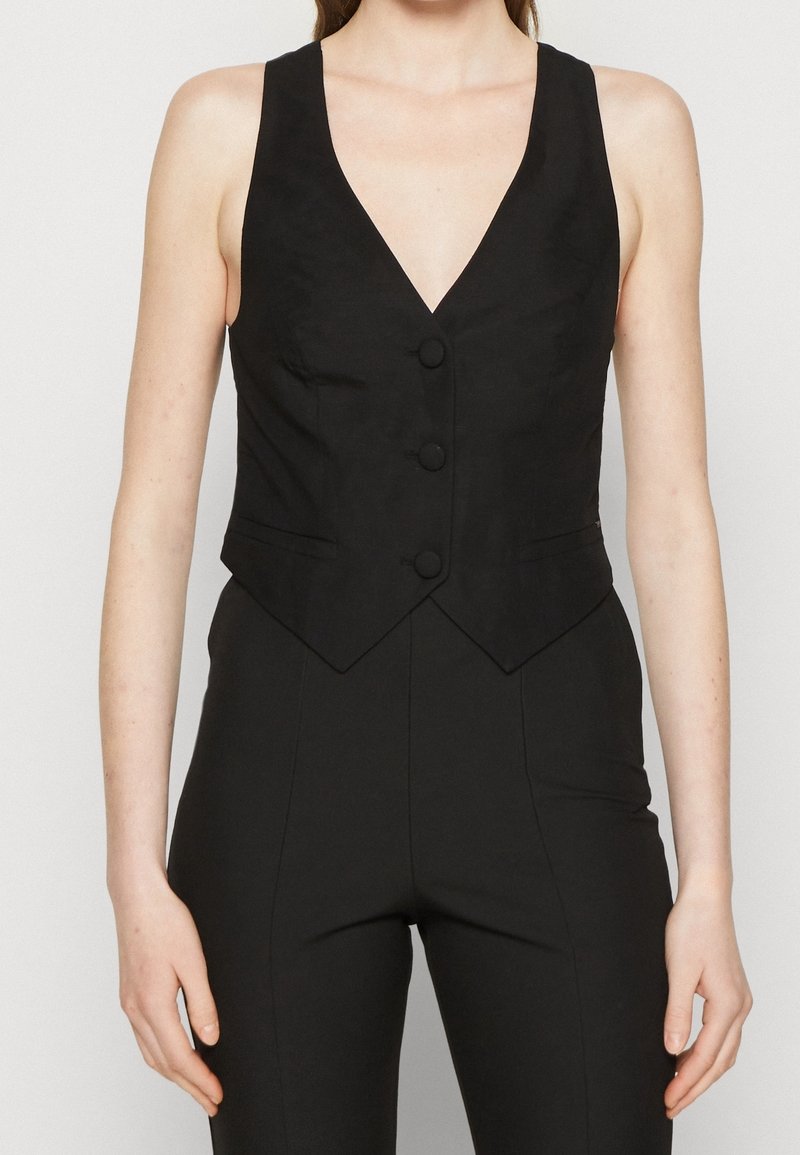 Svart väst med djup V-ringning, tre framknappar, strukturerad form och skräddarsydd passform. Tillverkad av ett slätt tyg utan synliga mönster.