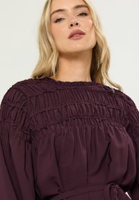 Abito viola scuro con dettagli smock, scollatura alta e vita legata. Realizzato in un morbido tessuto testurizzato con una silhouette fluida.