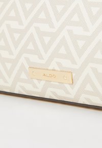 Kanvas skuldertaske i off-white med et geometrisk beige mønster. Har en guldfarvet mærkeplade og en sort accent i bunden.
