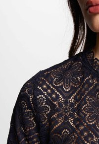 Top in pizzo blu navy caratterizzato da intricati motivi floreali, scollo alto e delicate rifiniture a scallop, che mette in risalto un tessuto trasparente e testurizzato.