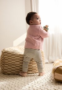 Lille barn iført en lyserød quiltet sweater og stribede leggings, der holder trælegetøj og står ved siden af en vævet puf på et tekstureret tæppe.