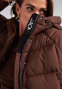 Brązowa kurtka puchowa z zamkiem błyskawicznym, z czarnymi detalami i logo. Pikowana tekstura, napy oraz regulowany kaptur.