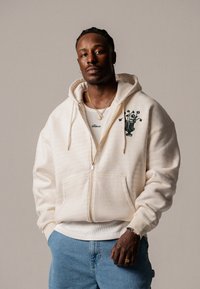 Krém színű, cipzáros kapucnis pulóver függőleges csíkokkal, zöld virágmintával hímzett. Fehér ujjatlan pólóval és farmernadrággal viselve, laza szabásban.