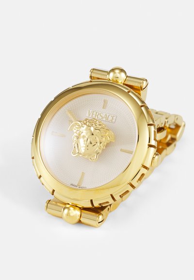 Zlaté náramkové hodinky s texturovaným bielym ciferníkom, ktorý obsahuje vyvýšené logo Medusa od Versace a zlaté akcenty. Dizajn s náramkom na články.