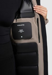 Beige puffjakke indvendig med sort polstring. Har et mærketeklæde, en lynlåslomme og glatte stofdetaljer.